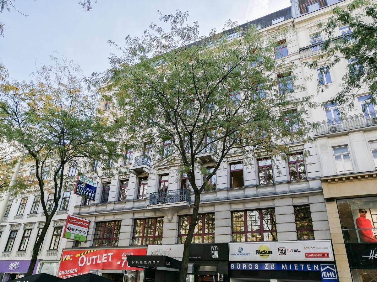 Ansicht der Gebäudefassade mit mehreren Etagen, Geschäften im Erdgeschoss und Bäumen davor in urbaner Umgebung.