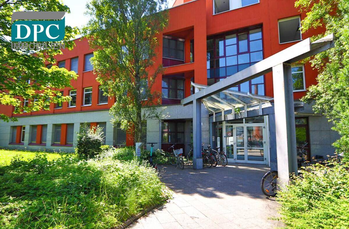 Eingangsbereich eines modernen Bürogebäudes mit Glasüberdachung und Fahrradständern, umgeben von üppigem Grün.