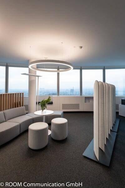 Büro-Loungebereich mit grauem Sofa, weißen Hockern und einem Raumteiler, umgeben von großen Fenstern.