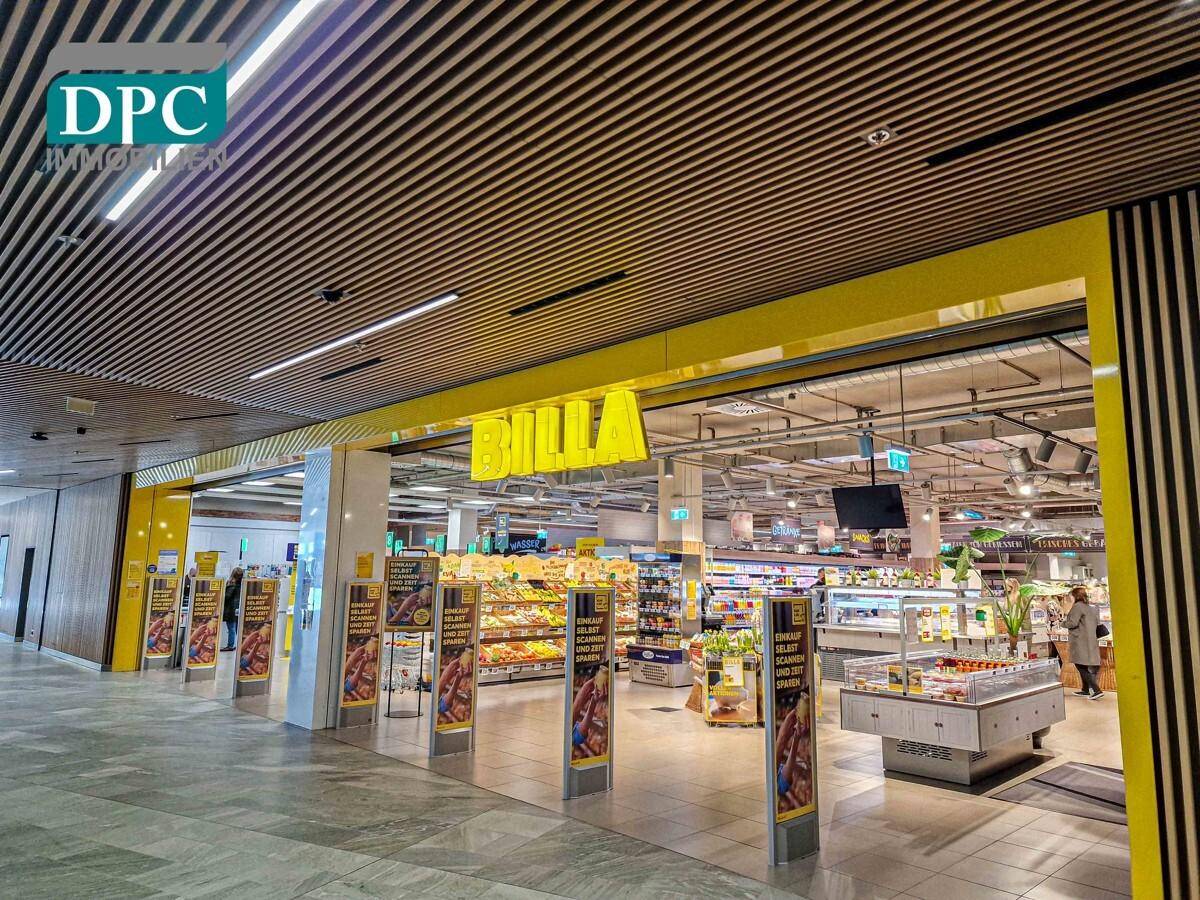 Eingangsbereich eines Supermarktes mit gelbem Logo, Regalen und Kunden, die einkaufen.