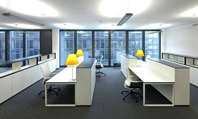 Modernes Großraumbüro mit mehreren Arbeitsplätzen, Teppichboden und großen Fenstern, die viel Tageslicht hereinlassen.