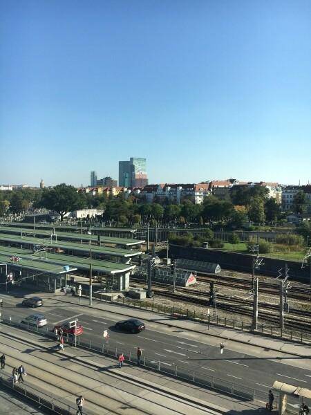 Weitläufiger Blick über eine Stadtlandschaft mit Bahngleisen, einer Straße und umliegenden Gebäuden unter blauem Himmel.