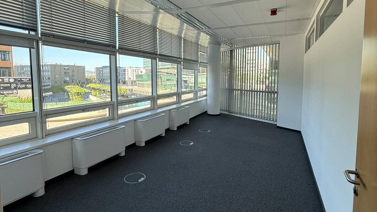 Helles, leeres Büro mit großen Fenstern, Teppichboden und Blick auf die Stadt.
