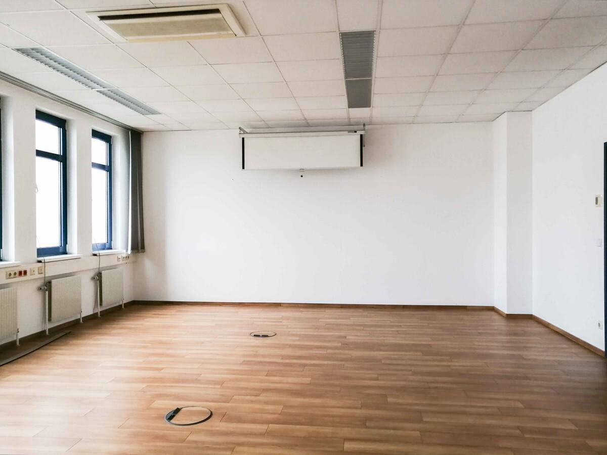 Leerer Büroraum mit Holzboden, weißen Wänden und ausfahrbarer Projektionsleinwand.