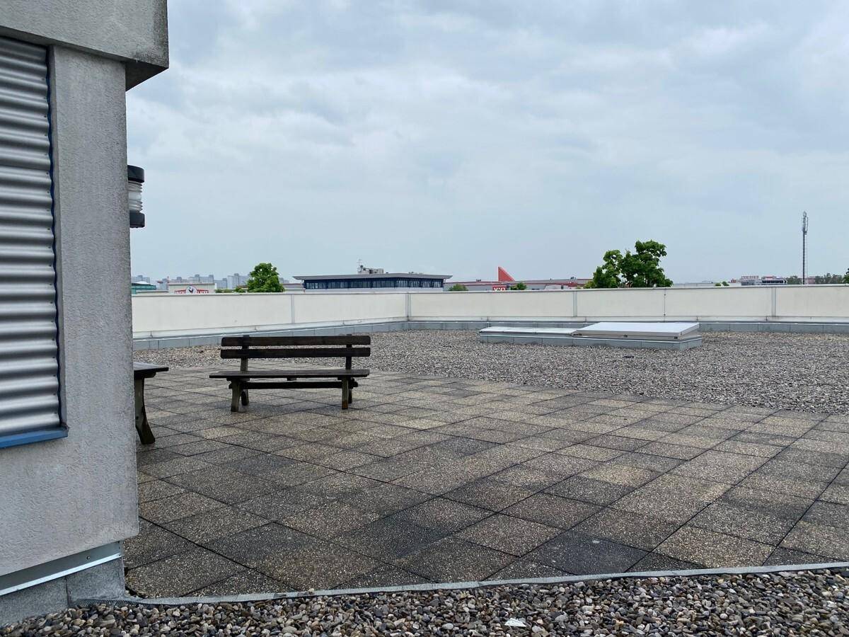 Dachterrasse mit Sitzbank und Kiesbereich, umgeben von einer niedrigen Brüstung unter bewölktem Himmel.