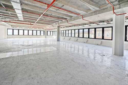 Heller, leerer Büroraum im Rohbau mit Betonboden, Säulen und einer Reihe von Fenstern entlang der Wand.