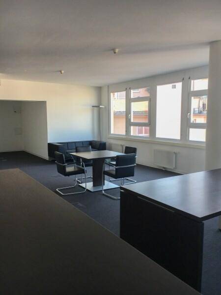 Moderner Bürobereich mit Sitzgelegenheiten, einem Besprechungstisch und großen Fenstern, die viel Tageslicht hereinlassen.