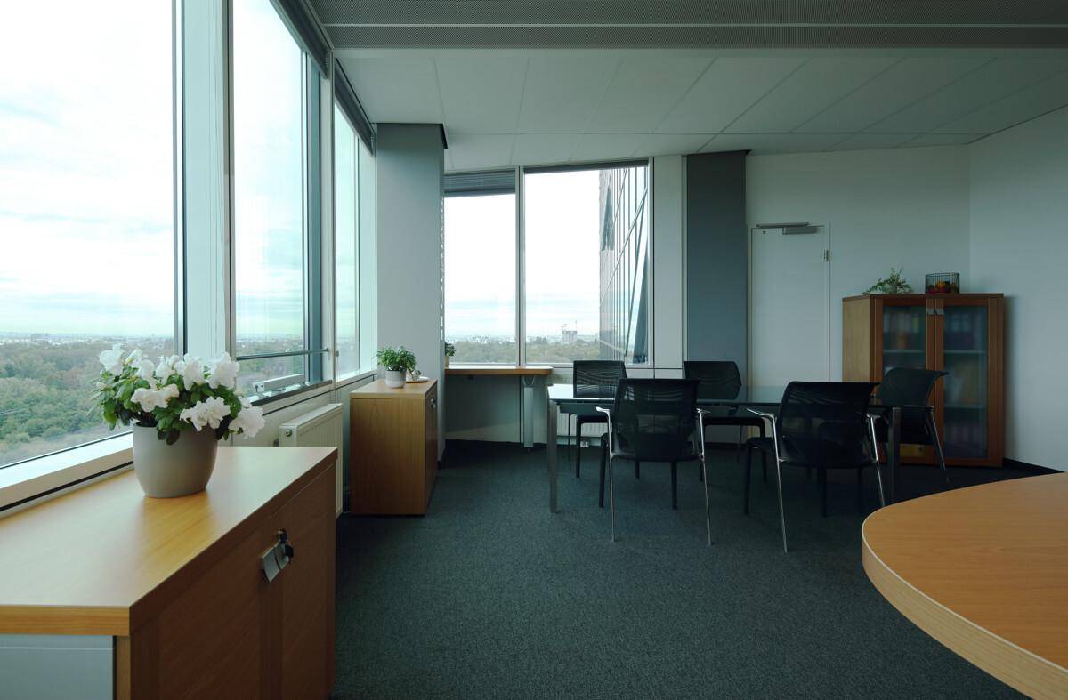 Helles Büro mit Schreibtischen, Schränken und großen Fenstern, die viel Tageslicht hereinlassen.