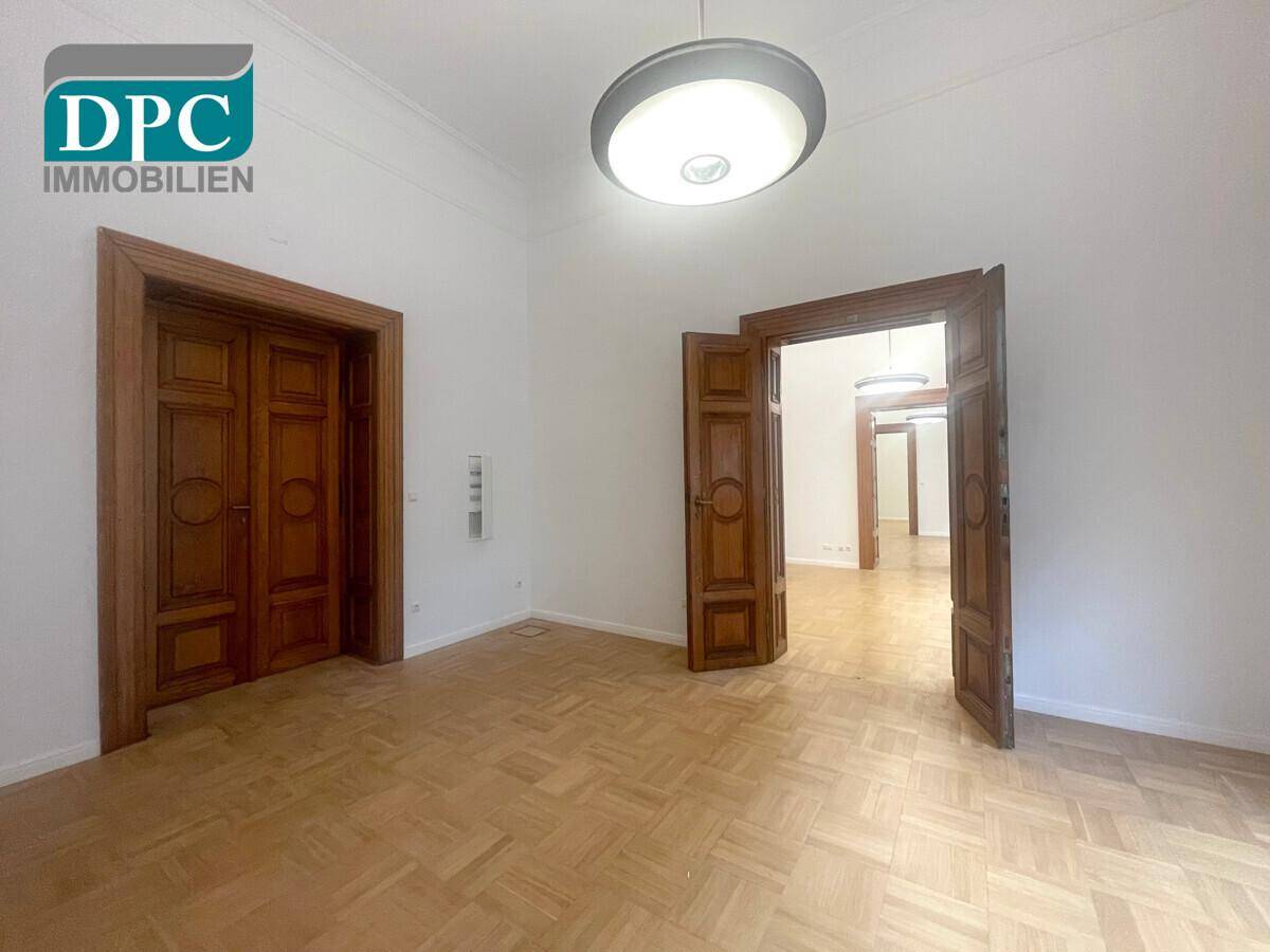 Heller Raum mit Fischgrätparkett und hohen Decken, mit Blick durch offene Holztüren in angrenzende Bereiche.