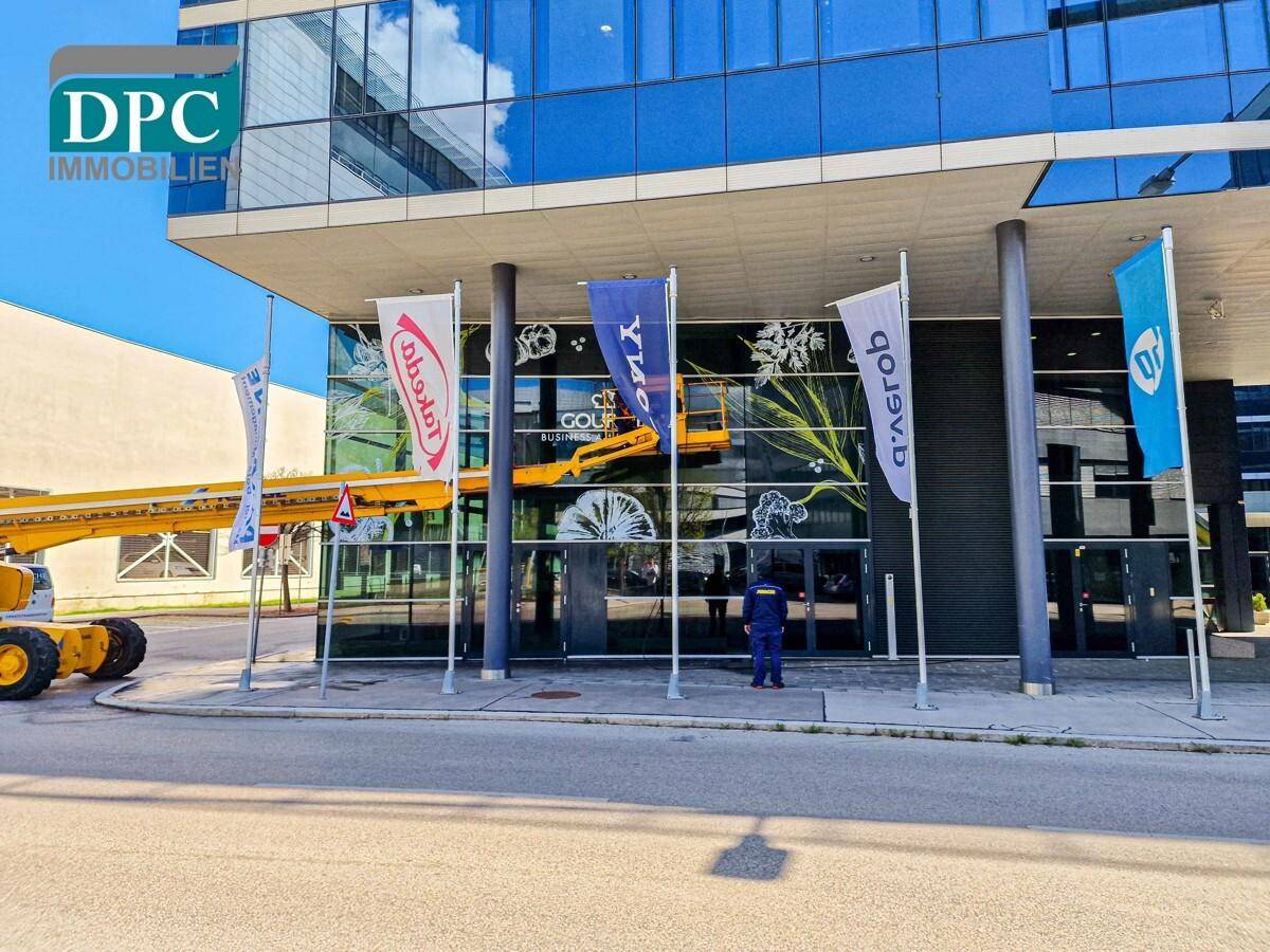 Modernes Geschäftsgebäude mit Glasfassade und Fahnen vor dem Eingangsbereich an einem sonnigen Tag.