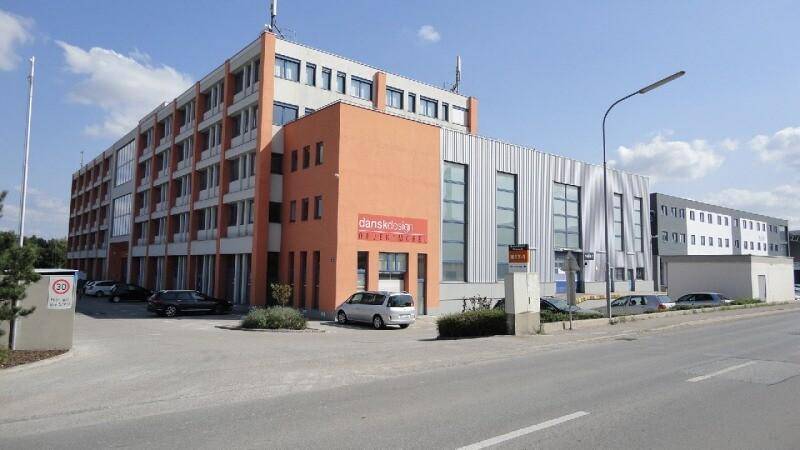 Modernes Geschäftsgebäude mit orangefarbenen und grauen Fassadenelementen sowie Parkplätzen im Vordergrund.