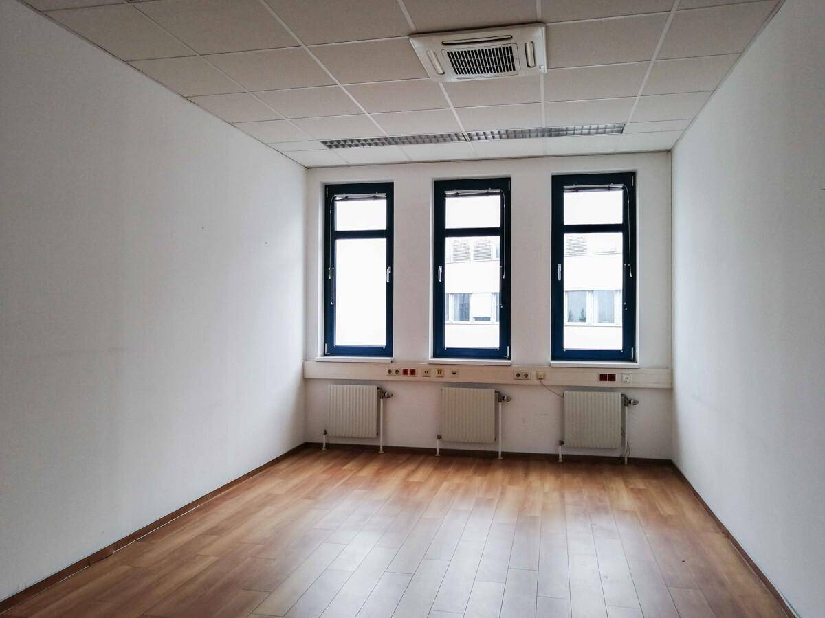 Leeres Büro mit Holzboden, weißen Wänden und drei Fenstern mit Heizkörpern darunter.