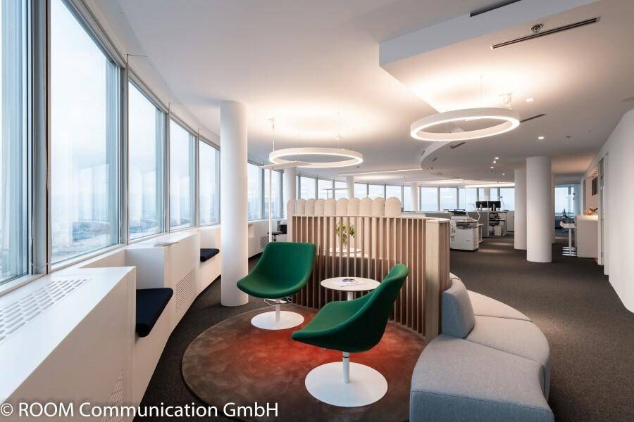 Moderner Büro-Loungebereich mit grünen Stühlen, geschwungenem Sofa und hölzernem Raumteiler.