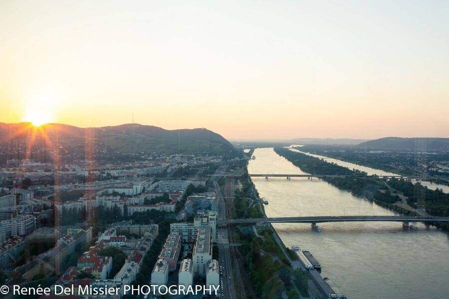 Spektakulärer Sonnenuntergang über der Stadt mit Fluss, Brücken und sanften Hügeln am Horizont.