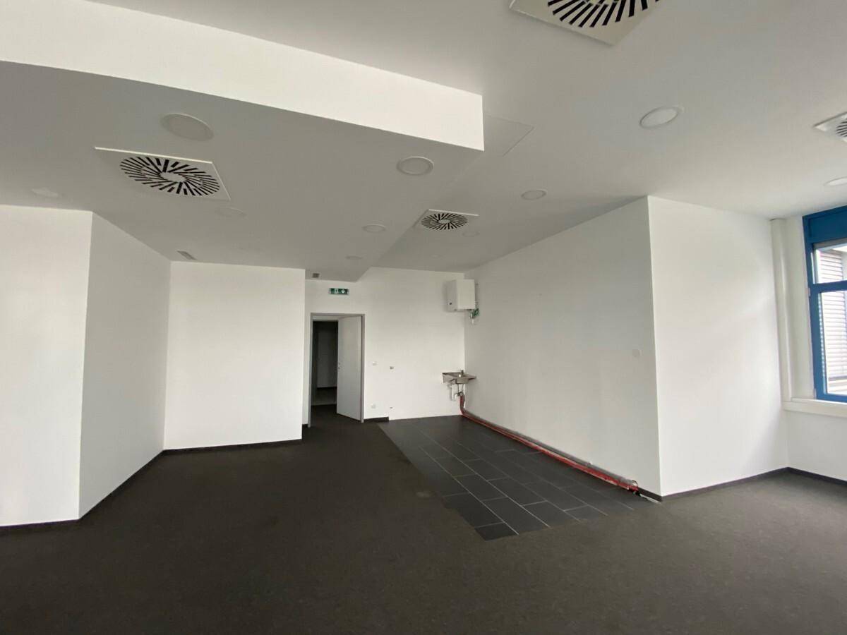 Bürobereich mit dunklem Boden, weiß gestrichenen Wänden, einem kleinen Waschbecken und Zugang zu weiteren Räumen.