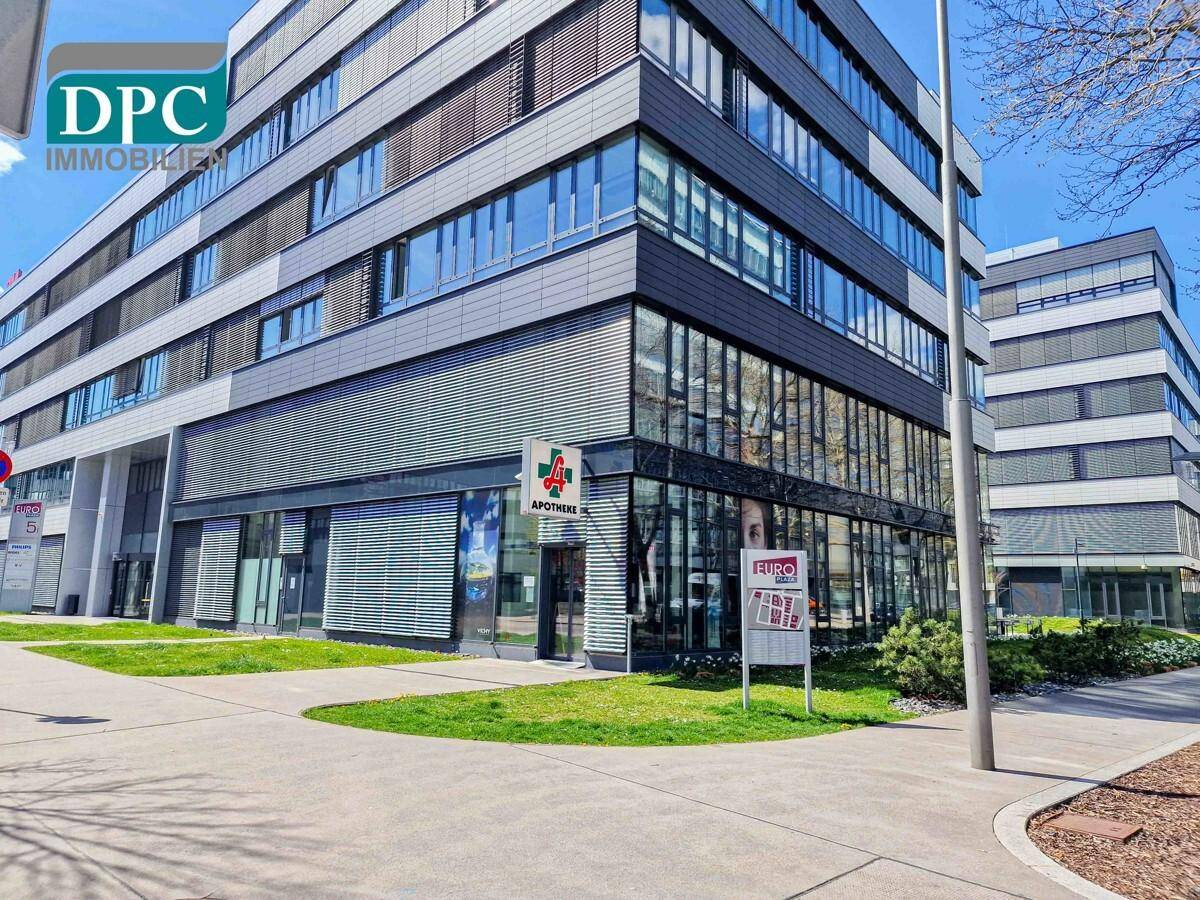 Modernes Geschäftsgebäude mit Apotheke im Erdgeschoss und großen Fensterfronten an einer städtischen Straße.