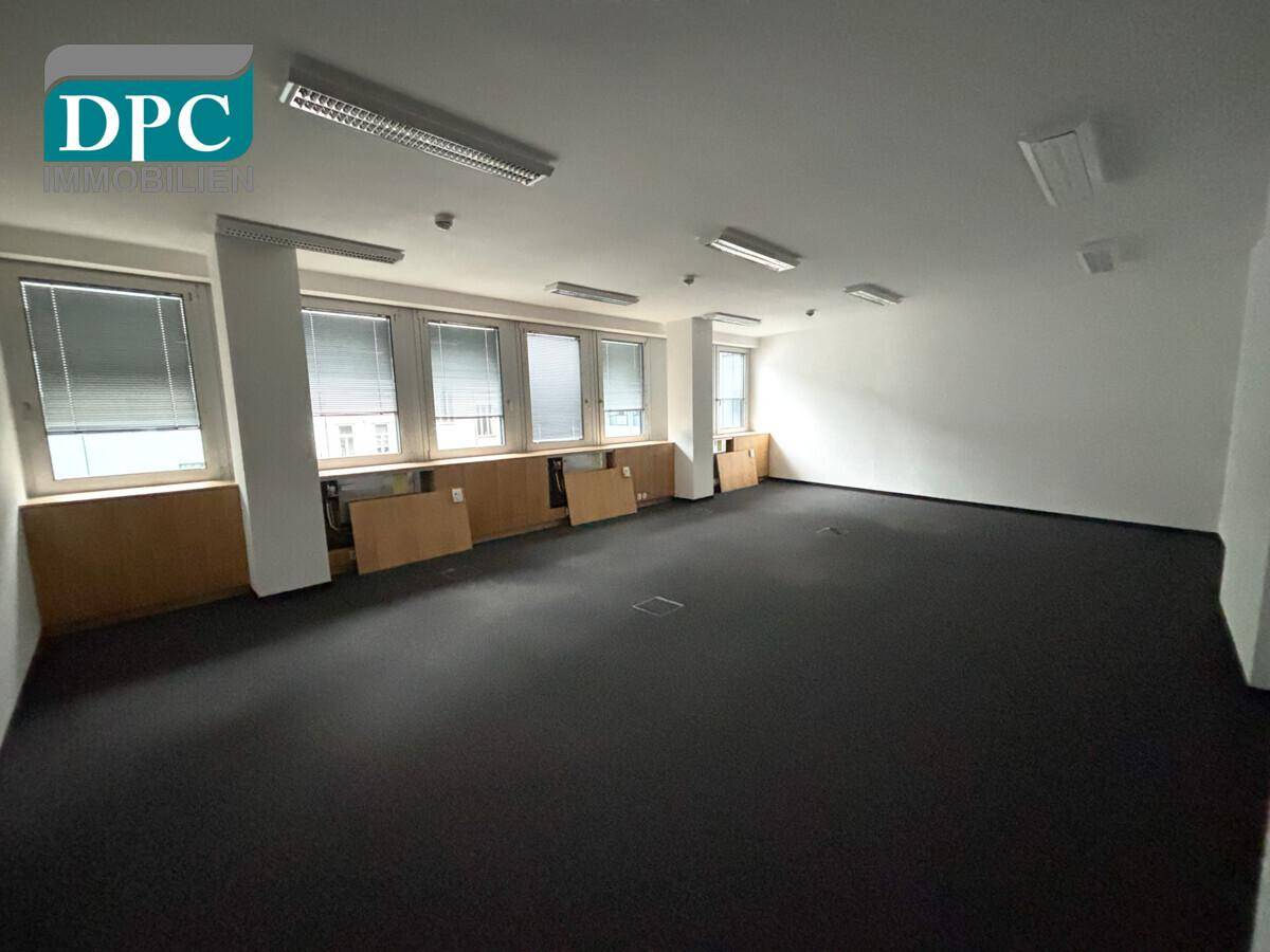 Geräumiges Büro mit Teppichboden, weißen Wänden und einer Fensterfront mit Jalousien.