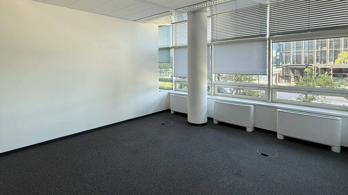 Geräumiges, leeres Büro mit Teppichboden, Säule und breiten Fenstern mit Jalousien.