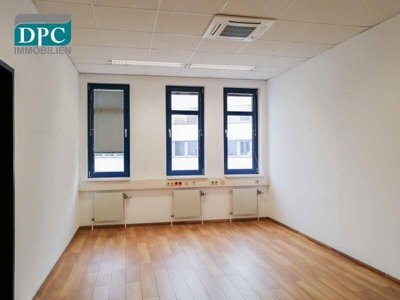 Leeres Büro mit Holzboden, weißen Wänden und drei Fenstern mit Heizkörpern darunter.