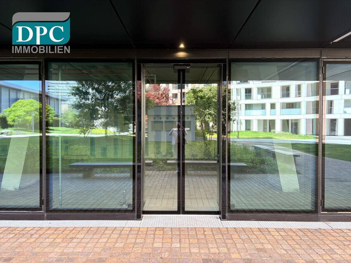 Moderne Glasfassade des Gebäudeeingangs, die den grünen Außenbereich und die umliegenden Gebäude widerspiegelt.