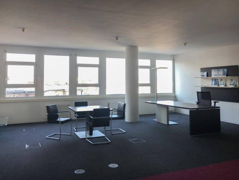 Geräumiger Bürobereich mit modernen Möbeln, einer Säule und großen Fenstern, die viel Tageslicht hereinlassen.