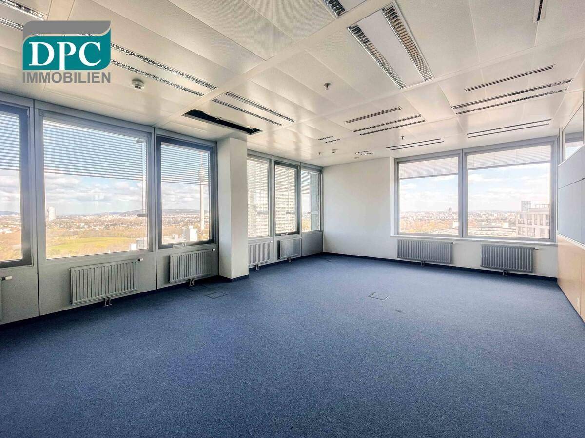 Geräumiges, leeres Büro mit blauem Teppichboden, großen Fenstern und weitem Blick über die Stadtlandschaft.