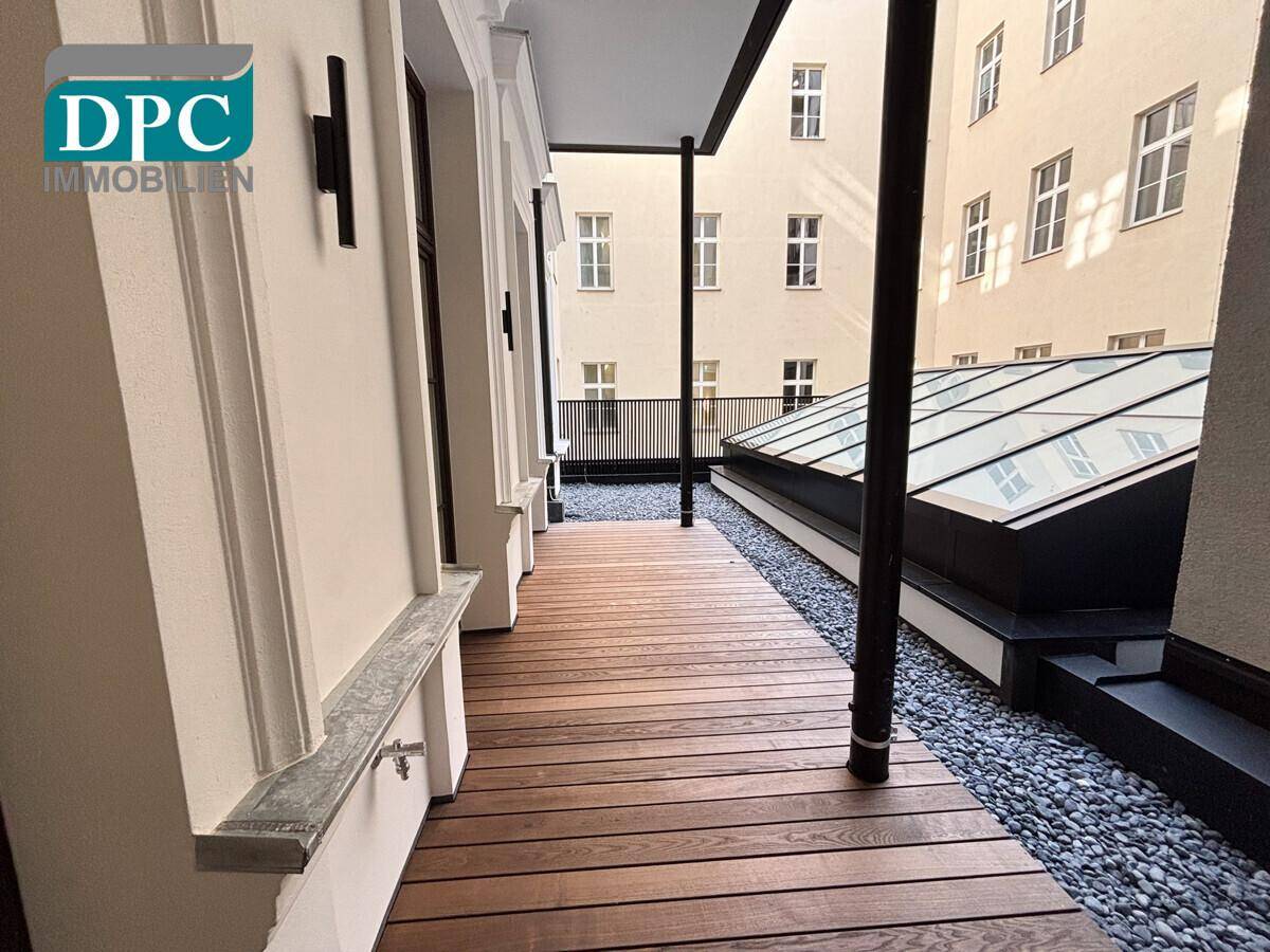 Außenbereich mit Holzplankenboden, Kiesbett und einem modernen Glasdach, umgeben von einer Gebäudefassade.