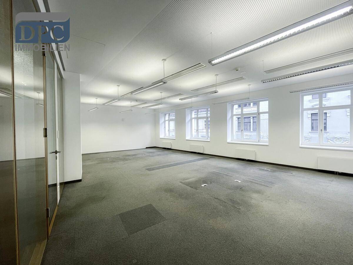 Geräumiger, leerer Büroraum mit grauem Teppichboden, weißen Wänden und großen Fenstern für viel Tageslicht.