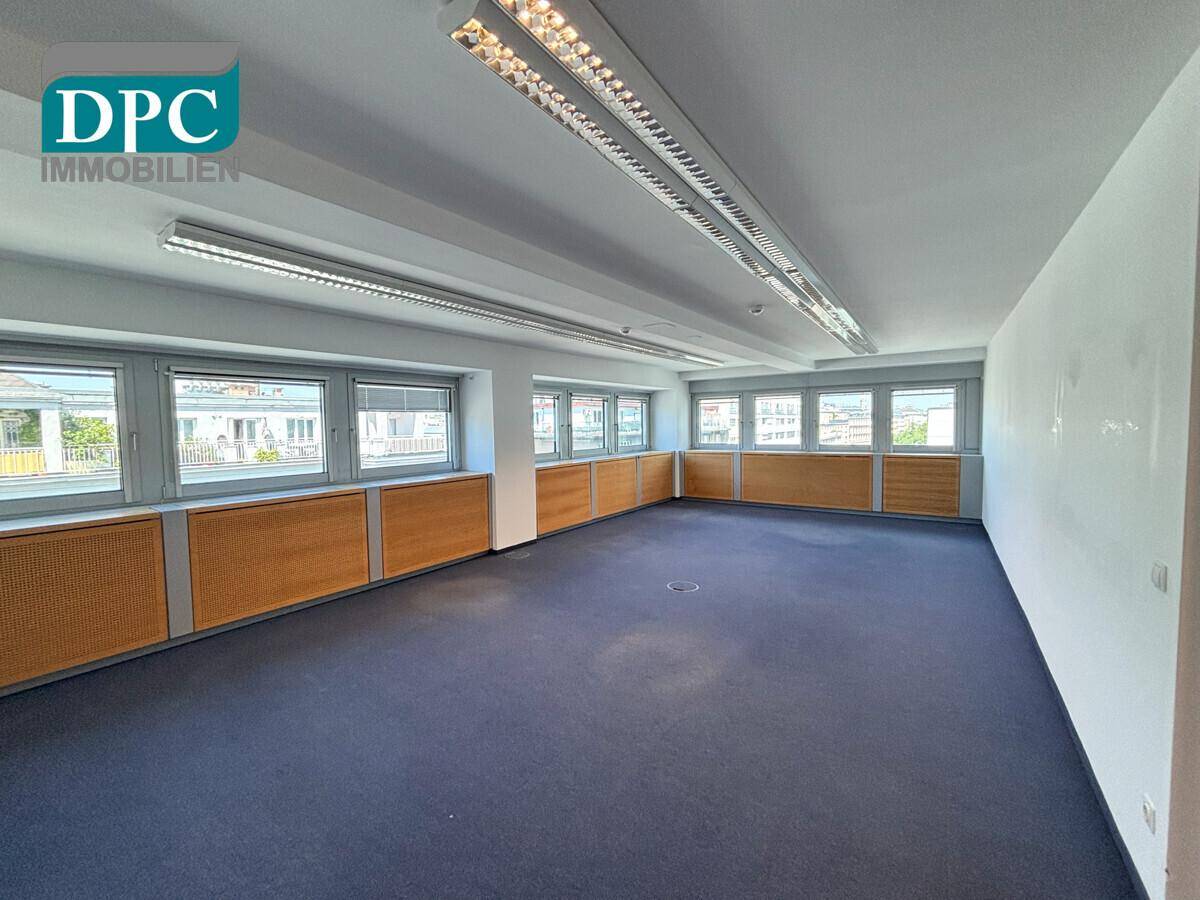 Geräumiges, leeres Büro mit blauem Teppichboden, weißen Wänden und großen Fenstern, ideal für gewerbliche Nutzung.