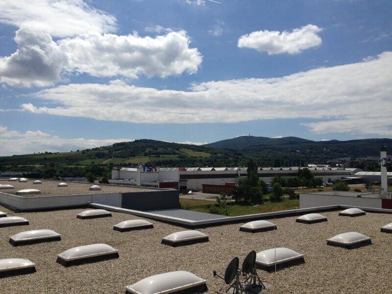 Blick von einem Dach mit vielen Oberlichtern auf eine grüne Hügellandschaft und weitere Gebäude in der Ferne.