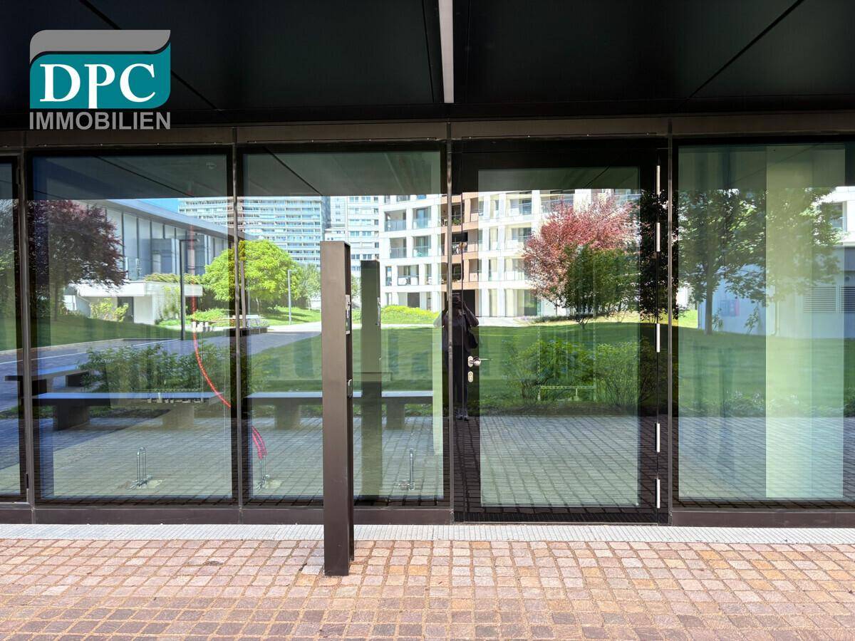 Moderne Glasfassade eines Gebäudes mit Blick auf eine grüne Außenanlage und weitere Gebäude.