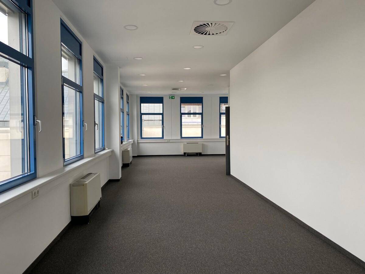 Heller Flur mit Teppichboden, Fenstern entlang einer Wand und Blick in einen größeren Bürobereich.