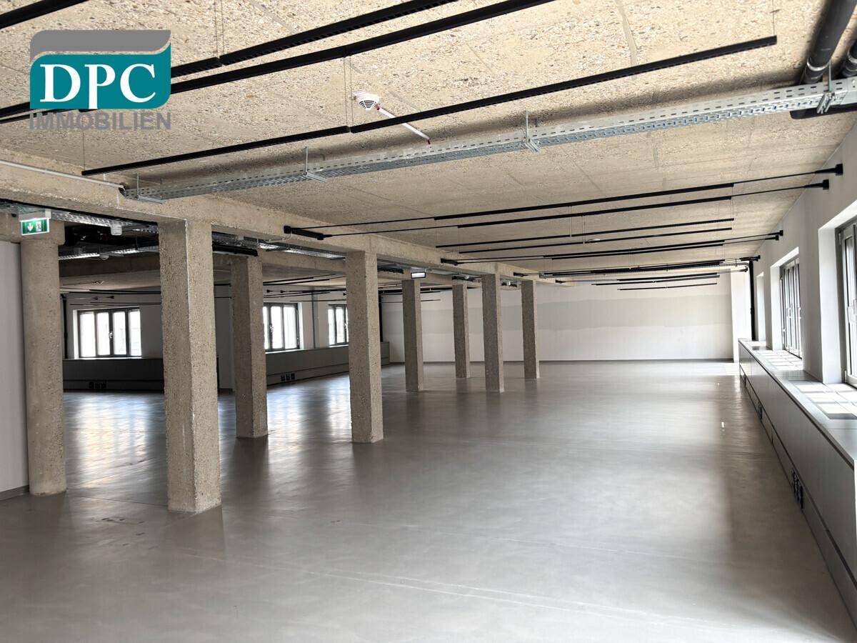 Weitläufiger, leerer Bürobereich mit Betonpfeilern, industrieller Decke und großen Fensterfronten.