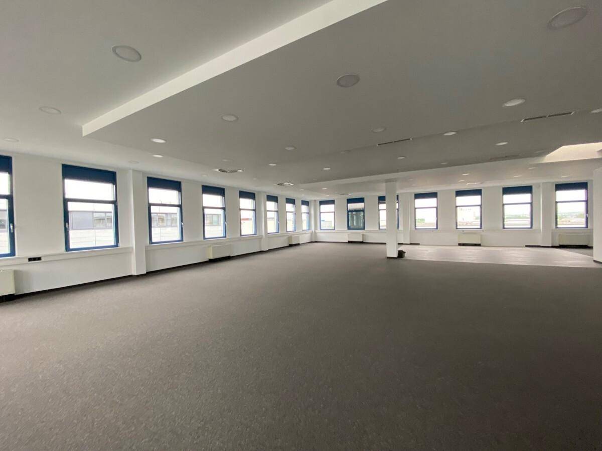 Heller, weitläufiger Büroraum mit durchgehender Fensterfront und dunklem Teppichboden, ideal für Großraumbüros.
