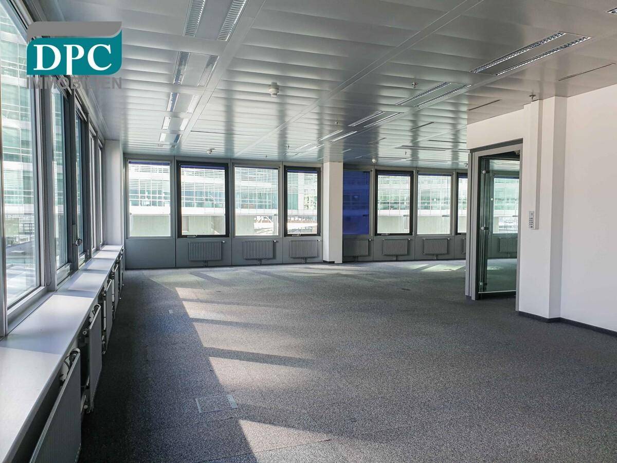 Geräumiger, leerer Büroraum mit Teppichboden, großen Fenstern und viel Tageslicht.