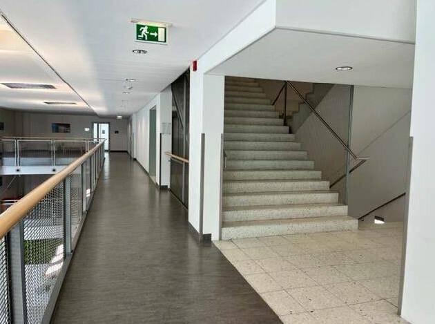 Heller Flur mit Treppenaufgang und Notausgangsschild, ausgestattet mit Handläufen und Fliesenboden.