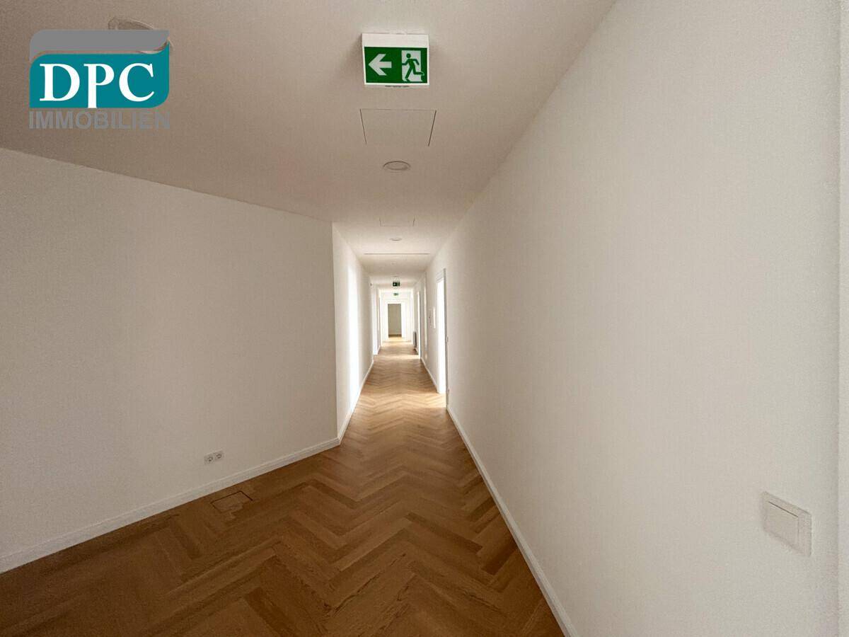 Langer, heller Flur mit Fischgrätparkett und Notausgangsschild an der Decke.