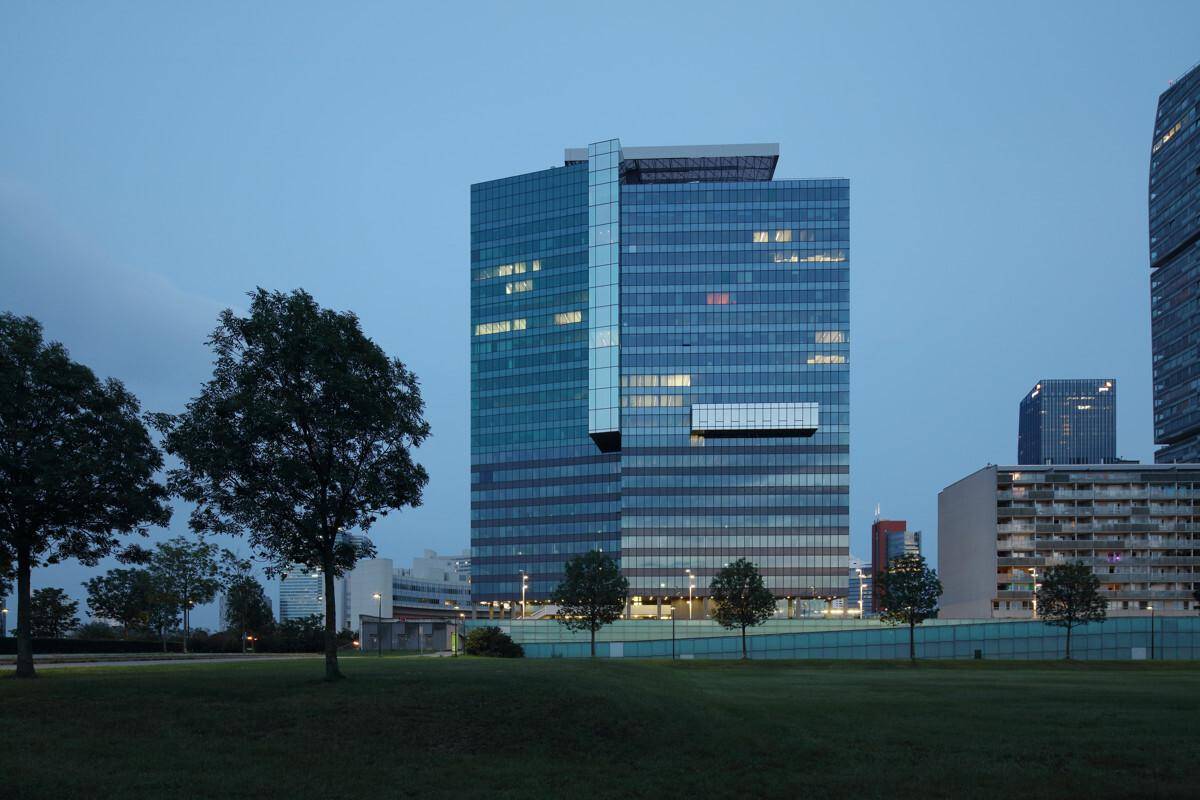 Modernes Hochhaus mit Glasfassade bei Dämmerung, umgeben von einer weitläufigen Grünfläche.