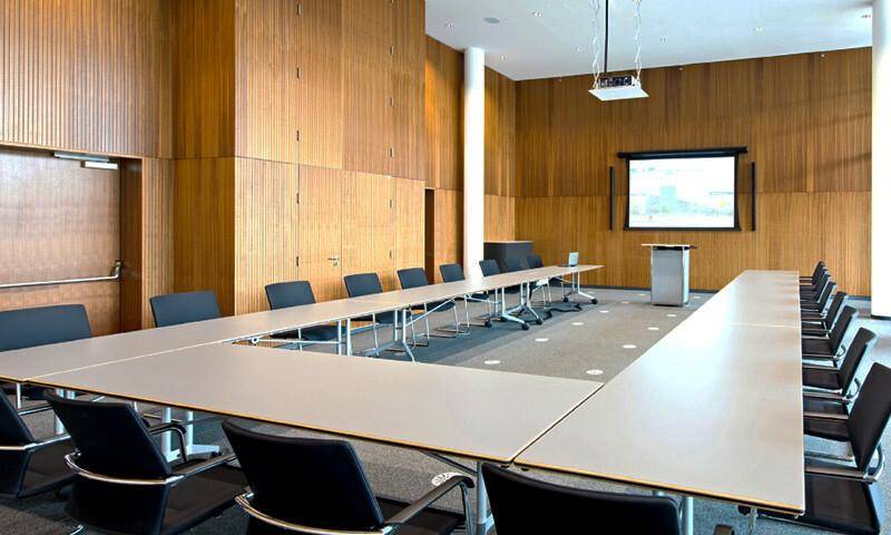 Großer Konferenzraum mit U-förmiger Tischordnung, Holzvertäfelung und einer Projektionsleinwand für Präsentationen.