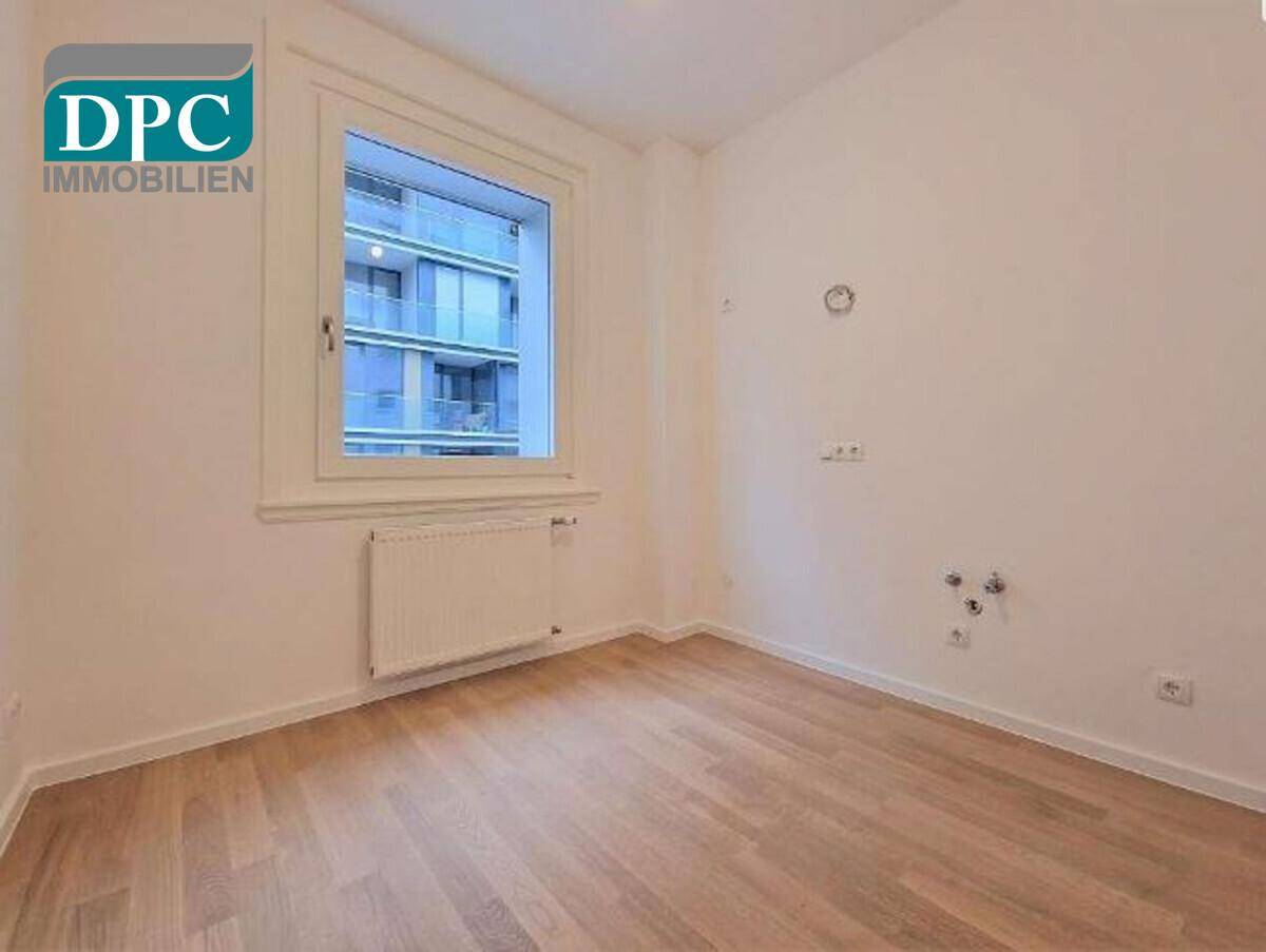 Leeres Zimmer mit Holzboden und einem großen Fenster, das einen Blick auf ein gegenüberliegendes Gebäude bietet.