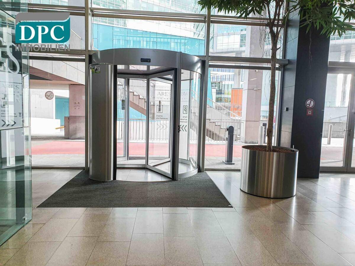 Heller Empfangsbereich mit Drehtür, Fliesenboden und einem dekorativen Baum in einem Pflanzkübel, einladend gestaltet.