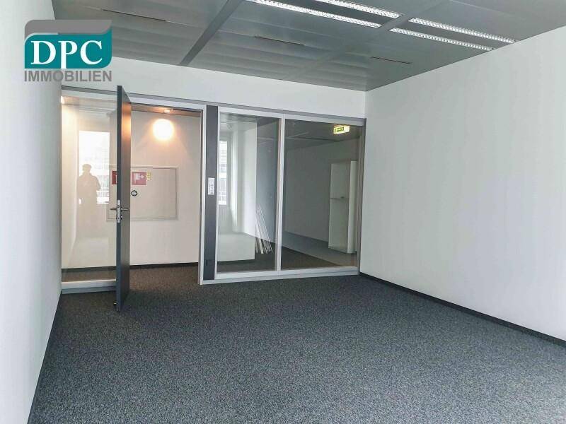 Bürofläche mit Teppichboden, weißen Wänden und Glastüren zu weiteren Räumen.