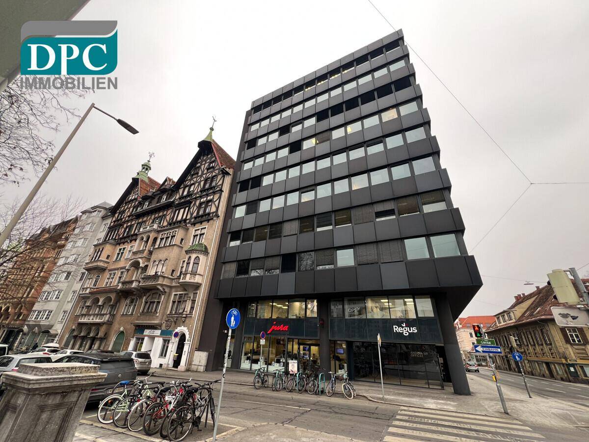 Moderne Geschäftsfassade mit dunklen Paneelen und großen Fenstern, gelegen an einer belebten Straßenecke neben einem Altbau.