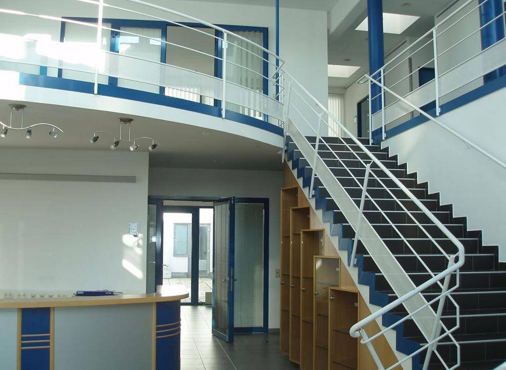 Heller Empfangsbereich mit markanter Treppe, blauen Akzenten und einer offenen Galerie im Obergeschoss.