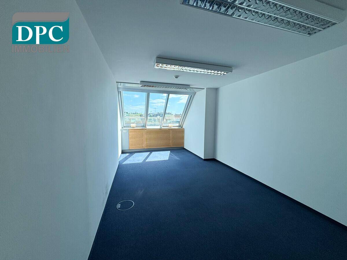 Helles Büro mit schrägen Fenstern, blauem Teppichboden und weiß gestrichenen Wänden, ideal für Arbeitsbereiche.