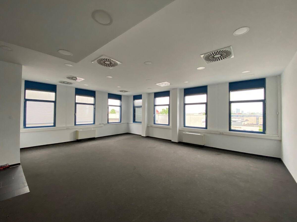 Heller Eckbereich eines Büros mit vielen Fenstern, dunklem Teppichboden und Deckenlüftungssystemen.