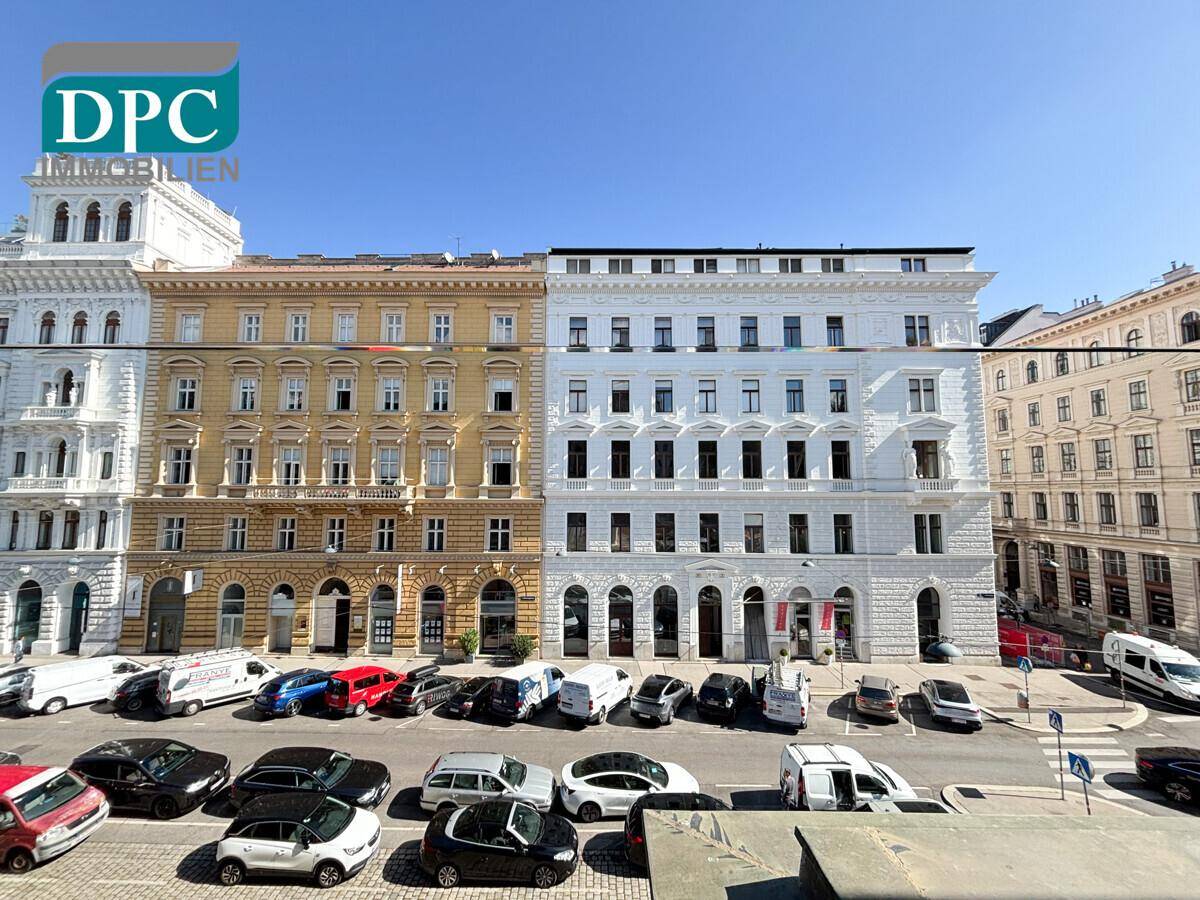 Historische Gebäudefronten entlang einer belebten Straße mit parkenden Autos unter blauem Himmel.