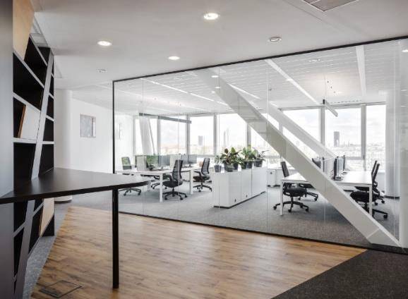 Modernes Büro mit Glaswänden, Holzboden und Teppichbereichen, ausgestattet mit Schreibtischen und Pflanzen.