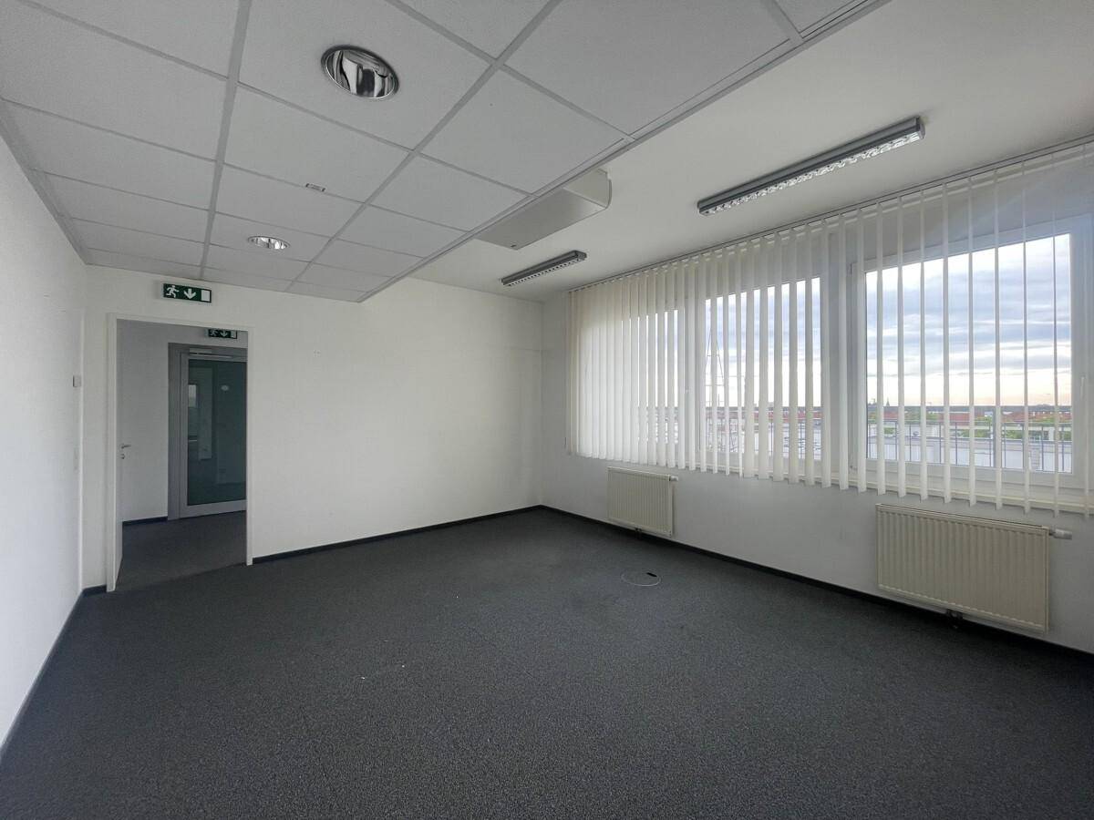 Helles Büro mit Teppichboden, weißen Wänden, Jalousien an den Fenstern und Deckenbeleuchtung.