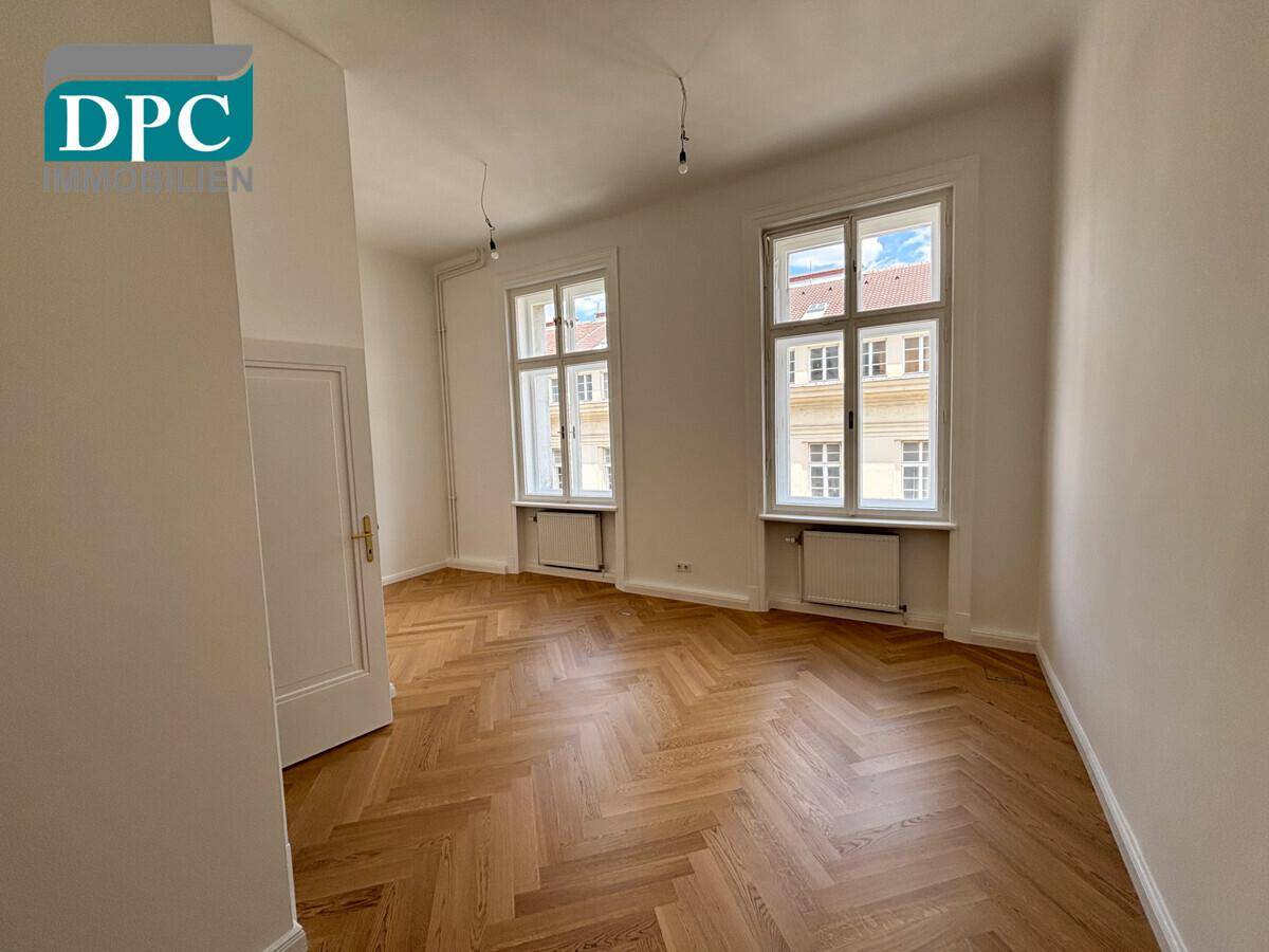Helles Zimmer mit Fischgrätparkett, hohen Fenstern und Blick auf die umliegenden Gebäude.